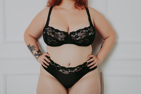 Full figured woman with plant tattoo on arm in black lace bra and panties with hands on hips